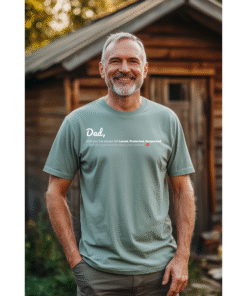 A smiling middle-aged man standing in front of his shed wearing a sage color tee shirt with a printed text design on the front