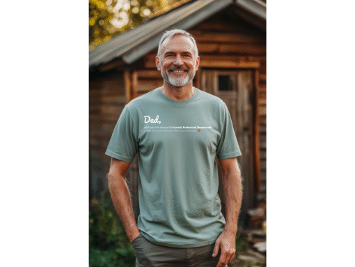 A smiling middle-aged man standing in front of his shed wearing a sage color tee shirt with a printed text design on the front